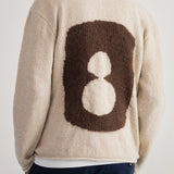 A person shown from the back wearing a cream-colored fuzzy knit sweater with a large brown and white graphic on the back, paired with dark navy pants, against a plain light background.