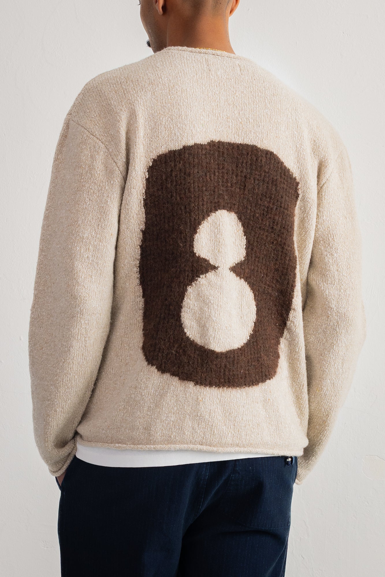 A person shown from the back wearing a cream-colored fuzzy knit sweater with a large brown and white graphic on the back, paired with dark navy pants, against a plain light background.
