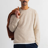 A man wearing a cream-colored knit crewneck sweater and dark navy trousers, holding a brown jacket over his shoulder against a plain white background.