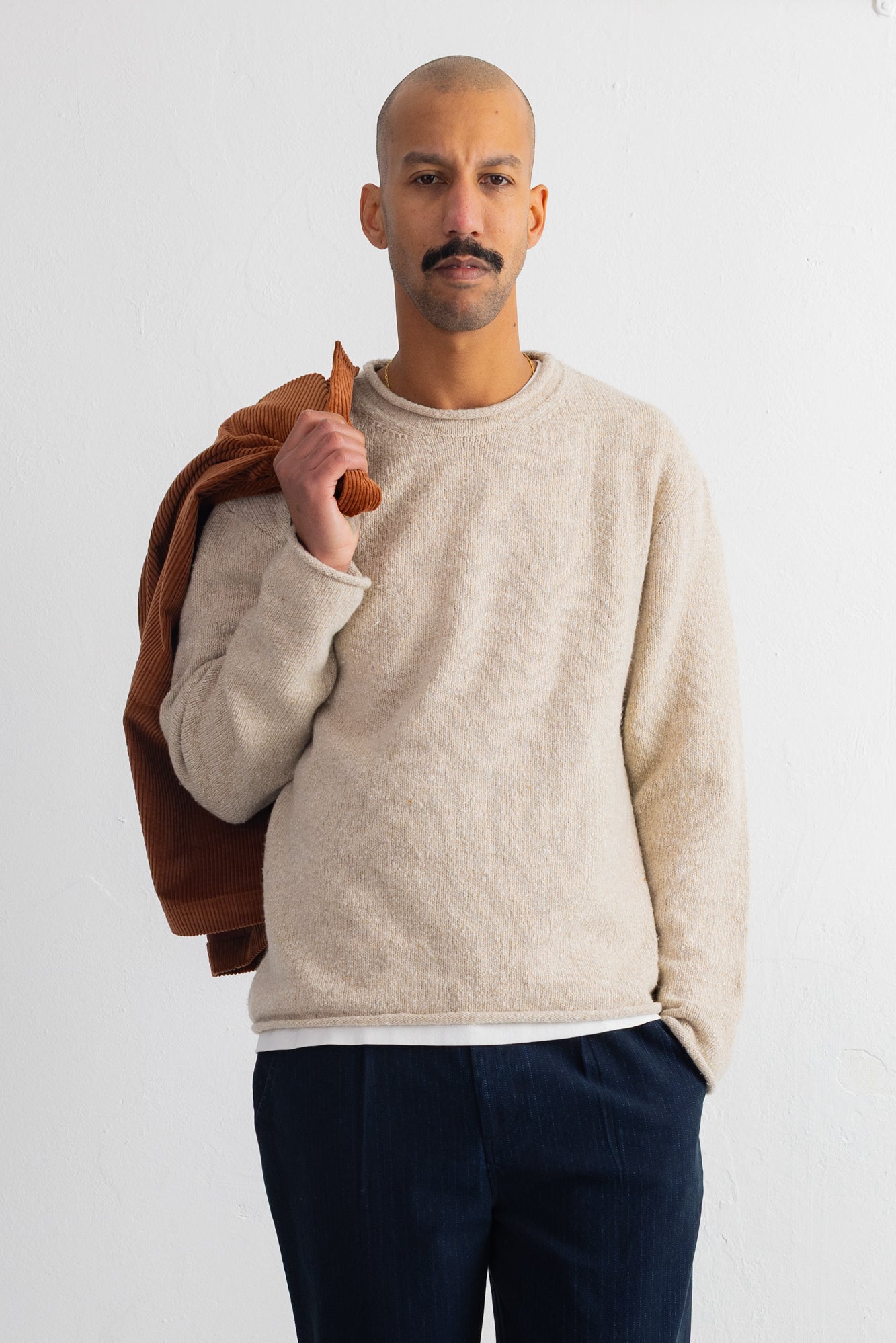 A man wearing a cream-colored knit crewneck sweater and dark navy trousers, holding a brown jacket over his shoulder against a plain white background.
