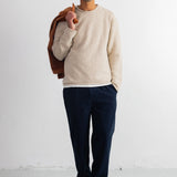 A man stands against a white studio wall wearing a cream knit crewneck sweater, dark navy trousers, black shoes, and holding a brown jacket over his shoulder.