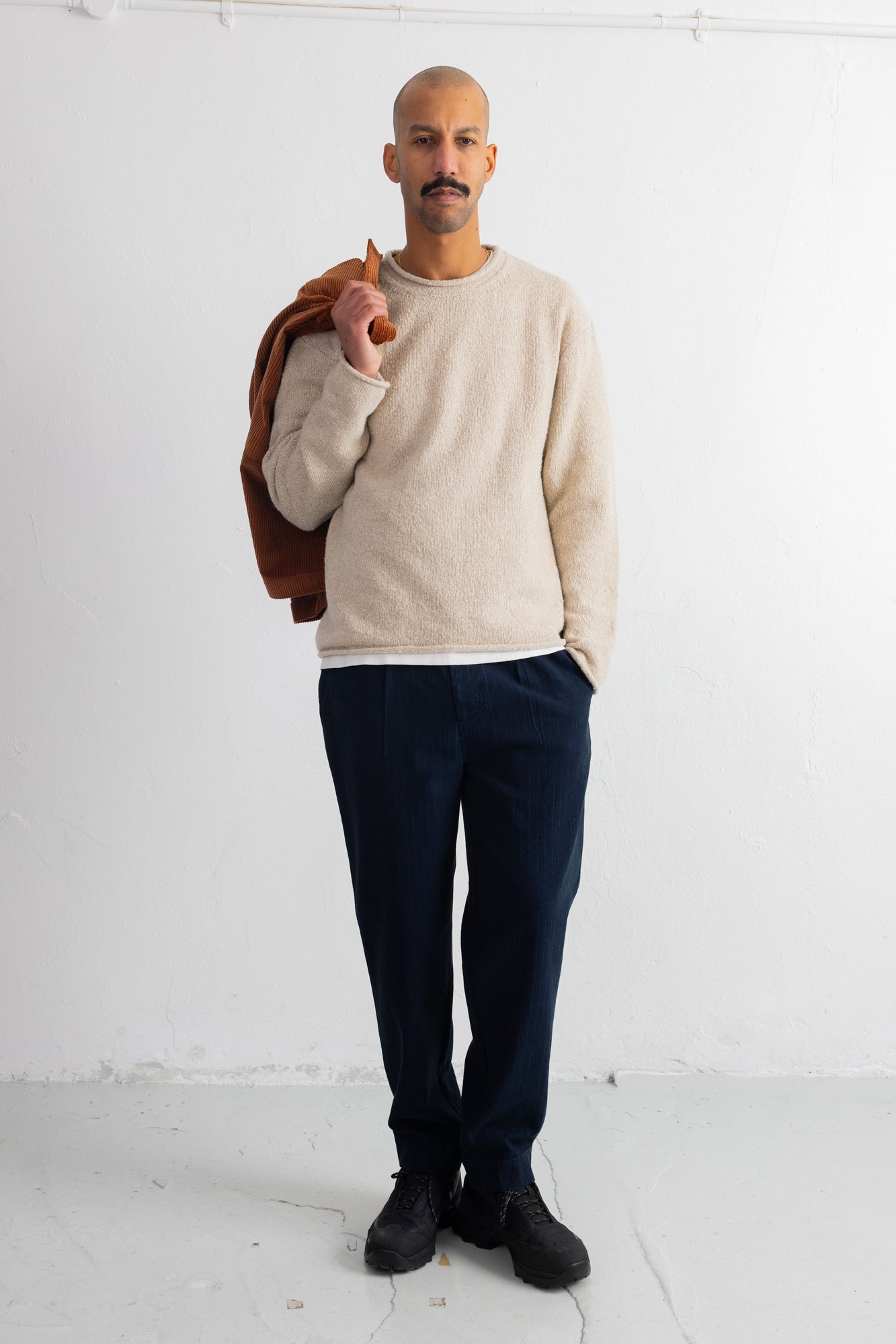 A man stands against a white studio wall wearing a cream knit crewneck sweater, dark navy trousers, black shoes, and holding a brown jacket over his shoulder.