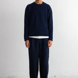 A man stands facing forward in a studio wearing a dark navy long-sleeve crewneck sweater over a white undershirt, matching navy pants, and black shoes against a plain white wall.