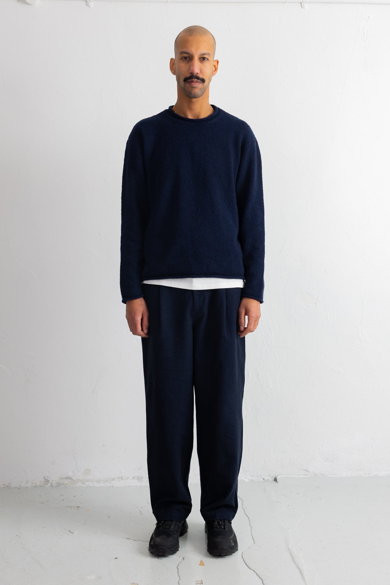 A man stands facing forward in a studio wearing a dark navy long-sleeve crewneck sweater over a white undershirt, matching navy pants, and black shoes against a plain white wall.