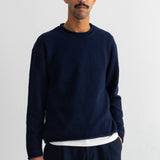 A man wearing a dark navy blue crewneck sweater over a white undershirt and matching navy pants, standing against a plain white wall.