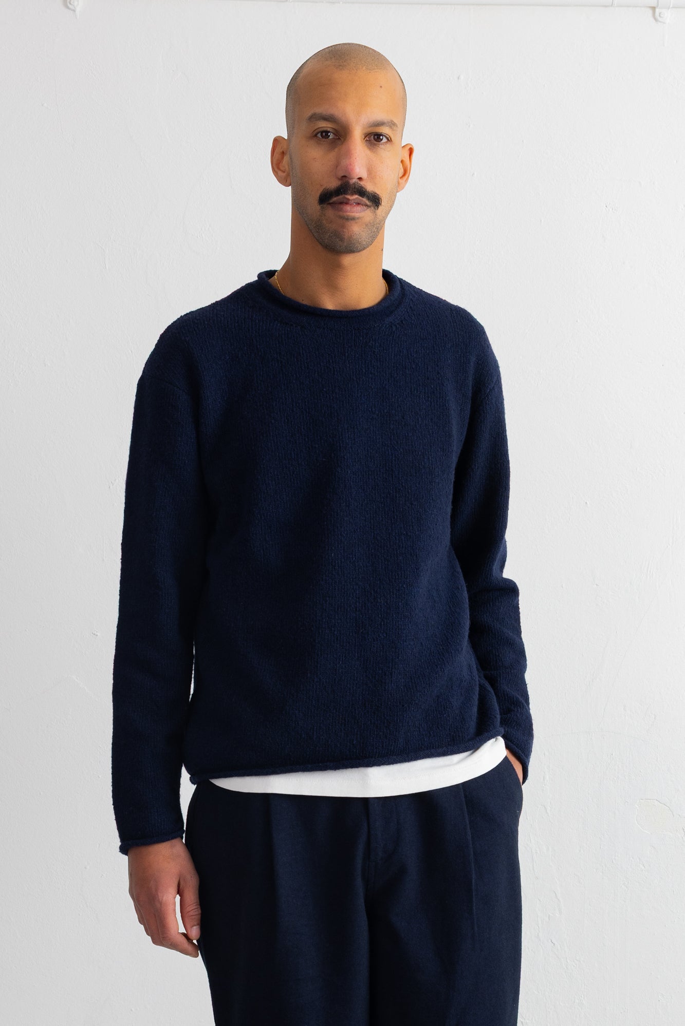 A man wearing a dark navy blue crewneck sweater over a white undershirt and matching navy pants, standing against a plain white wall.