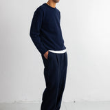 A man wearing a navy blue knit crewneck sweater and matching navy blue sweatpants, standing in profile against a white studio wall.