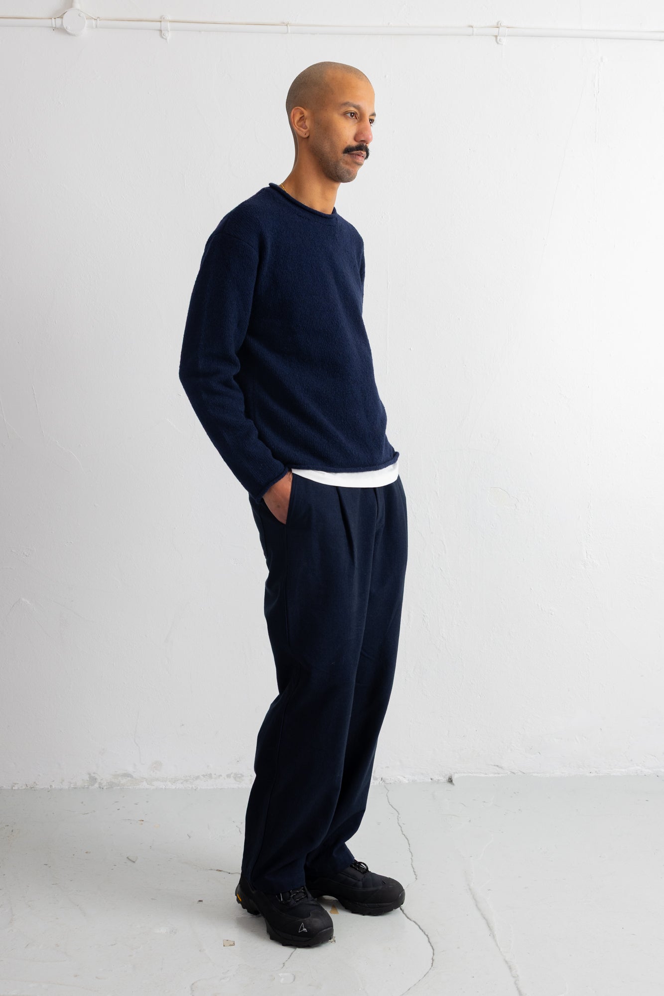 A man wearing a navy blue knit crewneck sweater and matching navy blue sweatpants, standing in profile against a white studio wall.