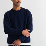 A man wearing a dark navy blue crewneck sweater with a contrasting white hem, paired with matching navy pants, standing against a plain white wall.