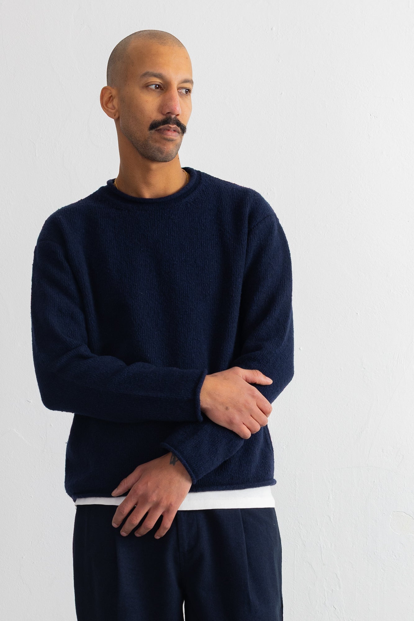 A man wearing a dark navy blue crewneck sweater with a contrasting white hem, paired with matching navy pants, standing against a plain white wall.