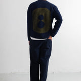 A man viewed from behind wearing a navy blue sweater with a large olive green textured number 8 on the back, paired with dark navy trousers and black shoes, standing against a white wall.