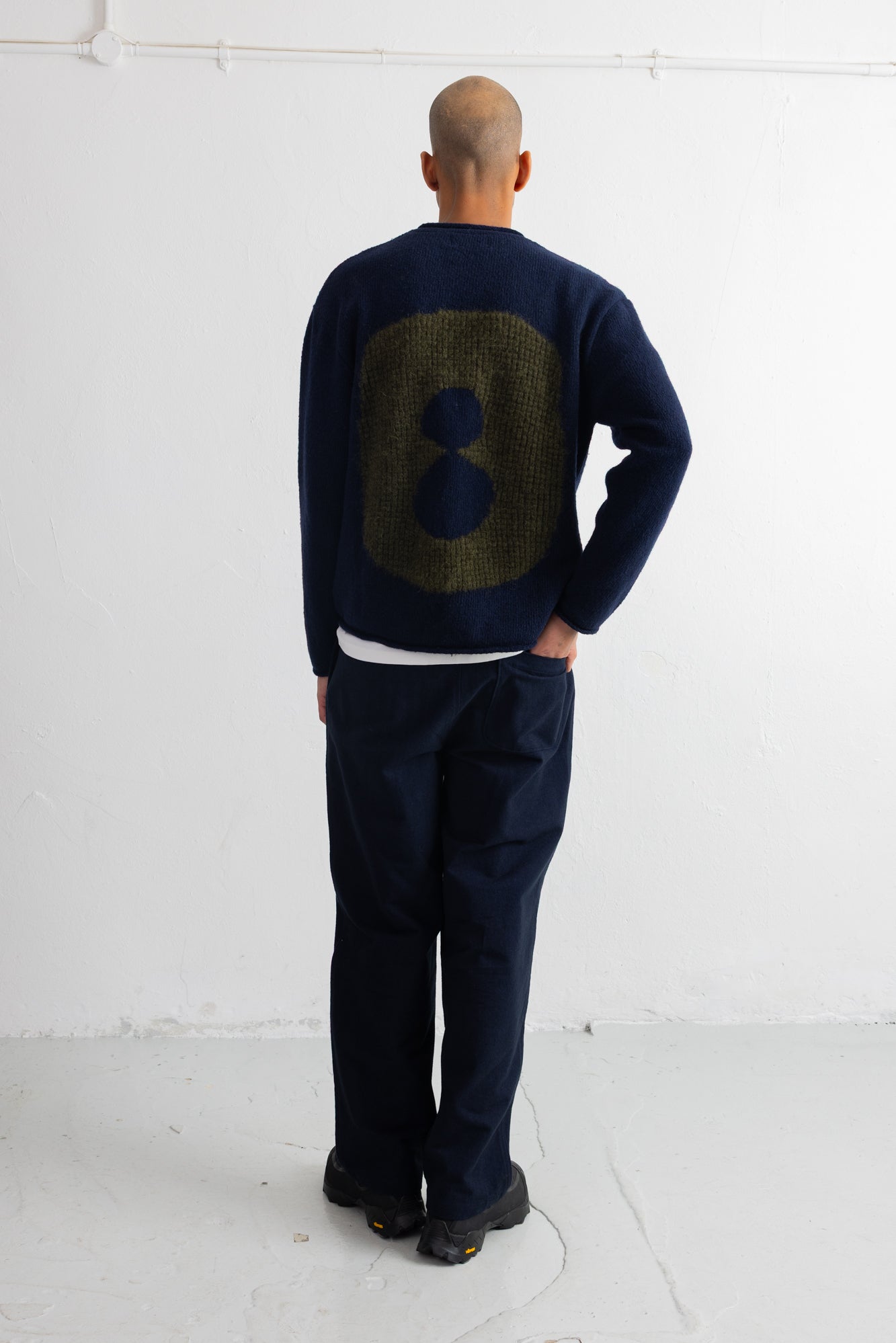 A man viewed from behind wearing a navy blue sweater with a large olive green textured number 8 on the back, paired with dark navy trousers and black shoes, standing against a white wall.