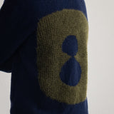 A close-up back view of a person wearing a navy blue knit sweater with a large olive green and navy blue abstract circular pattern on the back.
