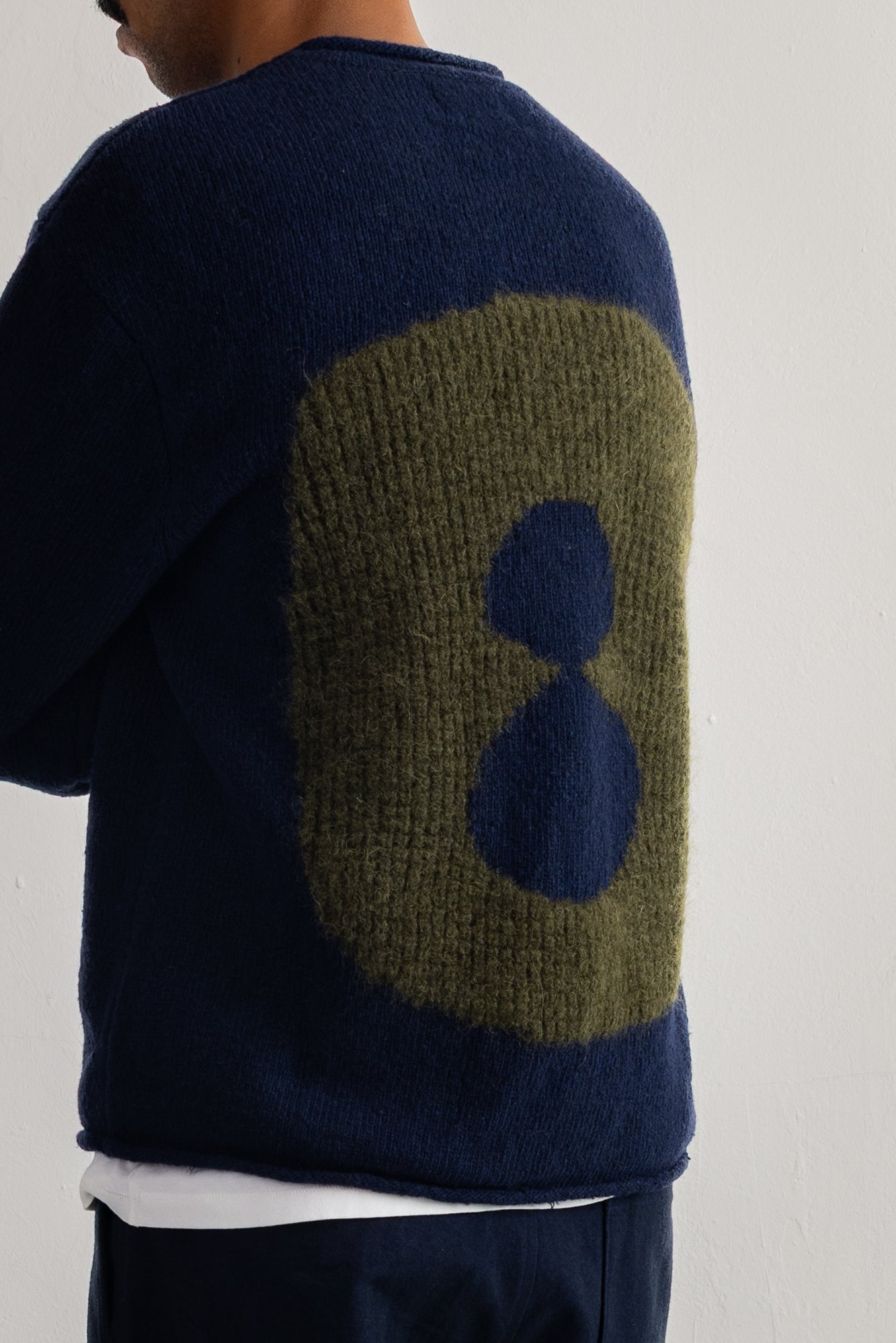 A close-up back view of a person wearing a navy blue knit sweater with a large olive green and navy blue abstract circular pattern on the back.