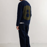 A man stands in profile wearing a navy blue knit sweater with a large olive green number 8 on the back, paired with dark navy trousers and black shoes, photographed against a plain white studio wall.