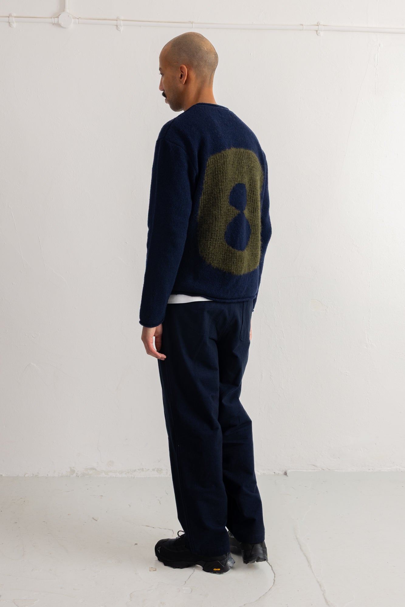 A man stands in profile wearing a navy blue knit sweater with a large olive green number 8 on the back, paired with dark navy trousers and black shoes, photographed against a plain white studio wall.