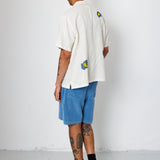 Person wearing a white shirt with colorful designs, blue shorts, and black sneakers against a white wall.