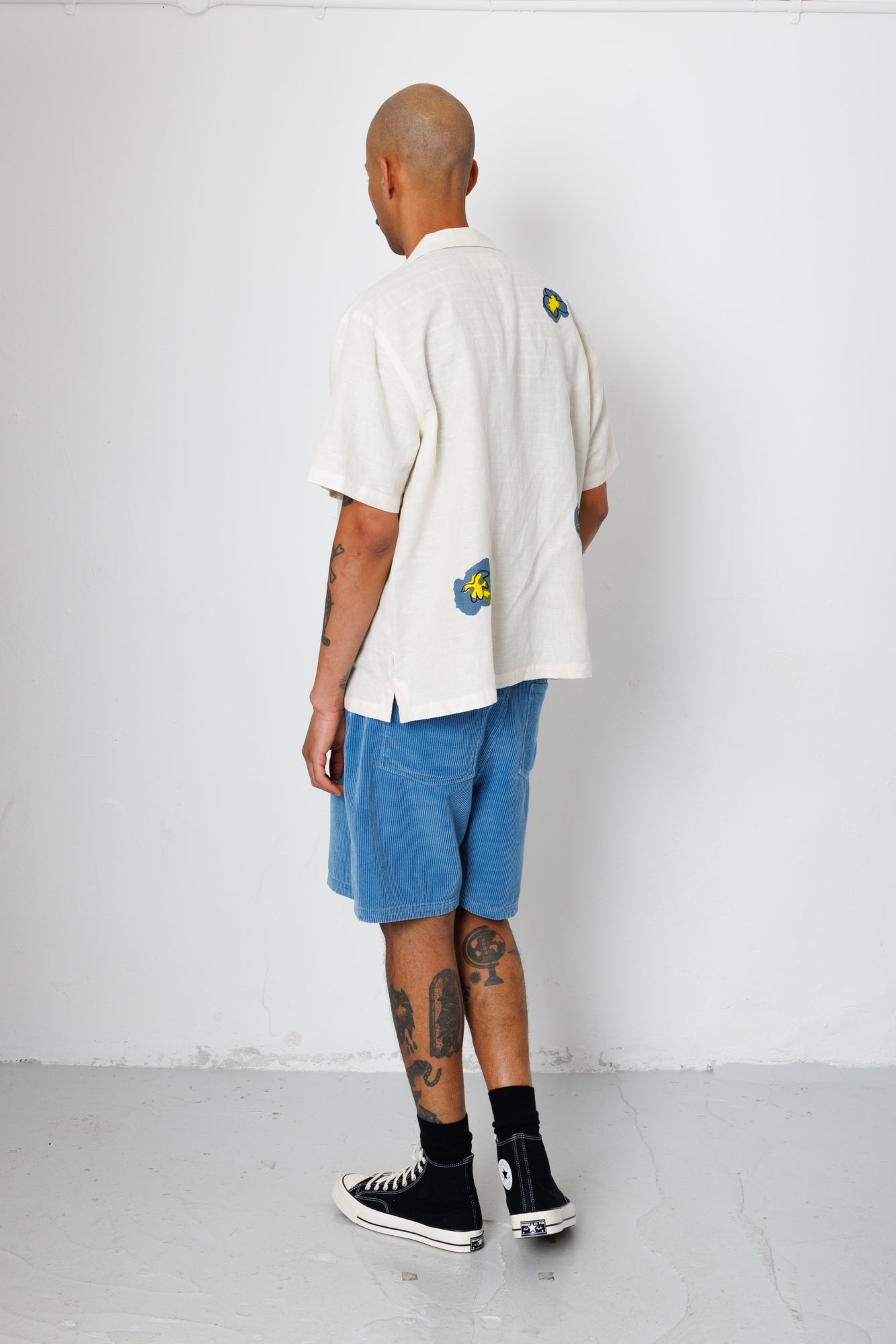 Person wearing a white shirt with colorful designs, blue shorts, and black sneakers against a white wall.