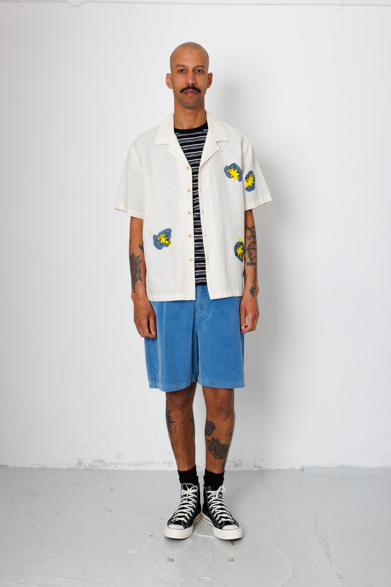 Man wearing a white shirt with colorful designs, blue shorts, and black sneakers against a white background