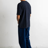 A man viewed from behind wearing a black short-sleeve t-shirt with thin blue horizontal stripes, dark blue jeans, and black canvas sneakers, standing against a white wall.