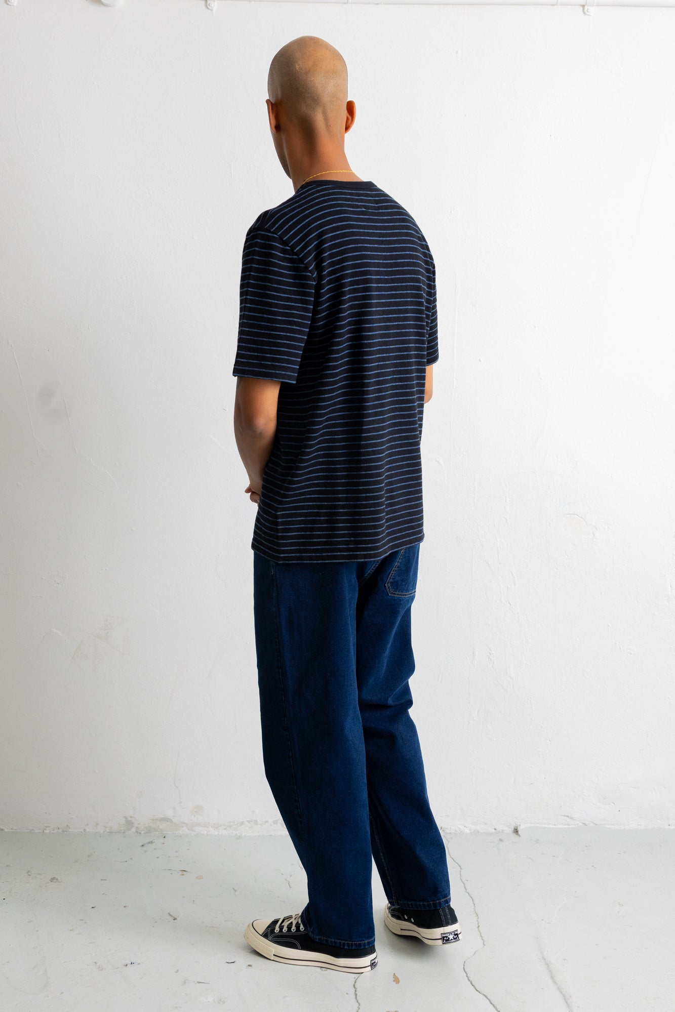 A man viewed from behind wearing a black short-sleeve t-shirt with thin blue horizontal stripes, dark blue jeans, and black canvas sneakers, standing against a white wall.