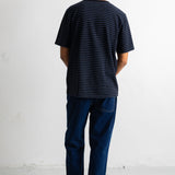 A man viewed from behind wearing a black short-sleeve t-shirt with thin horizontal blue stripes, dark blue jeans, and black-and-white sneakers, standing against a plain white wall.