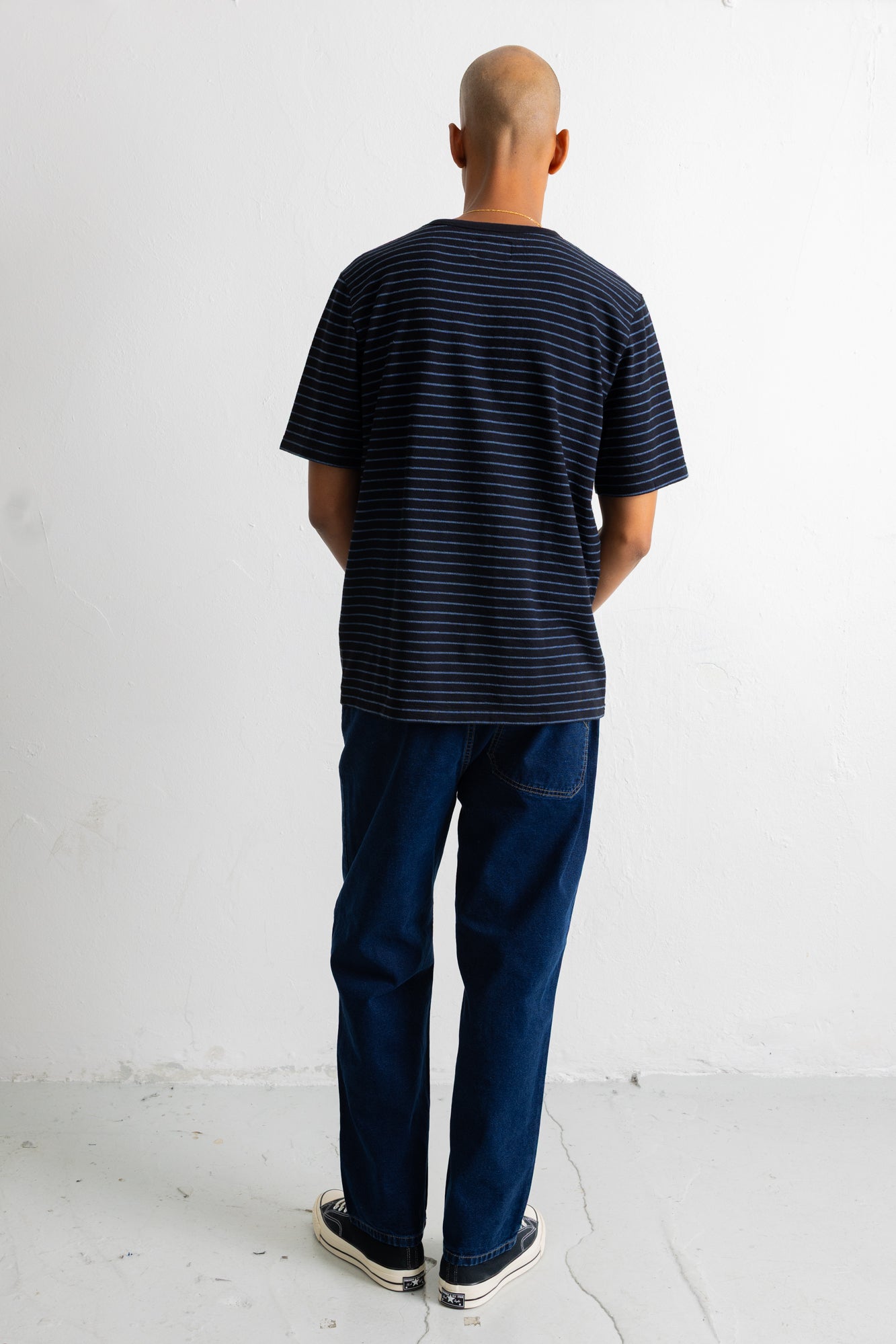 A man viewed from behind wearing a black short-sleeve t-shirt with thin horizontal blue stripes, dark blue jeans, and black-and-white sneakers, standing against a plain white wall.