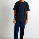 A man wearing a black short-sleeve t-shirt with thin horizontal blue stripes, dark blue jeans, and black-and-white sneakers, standing against a white studio wall.