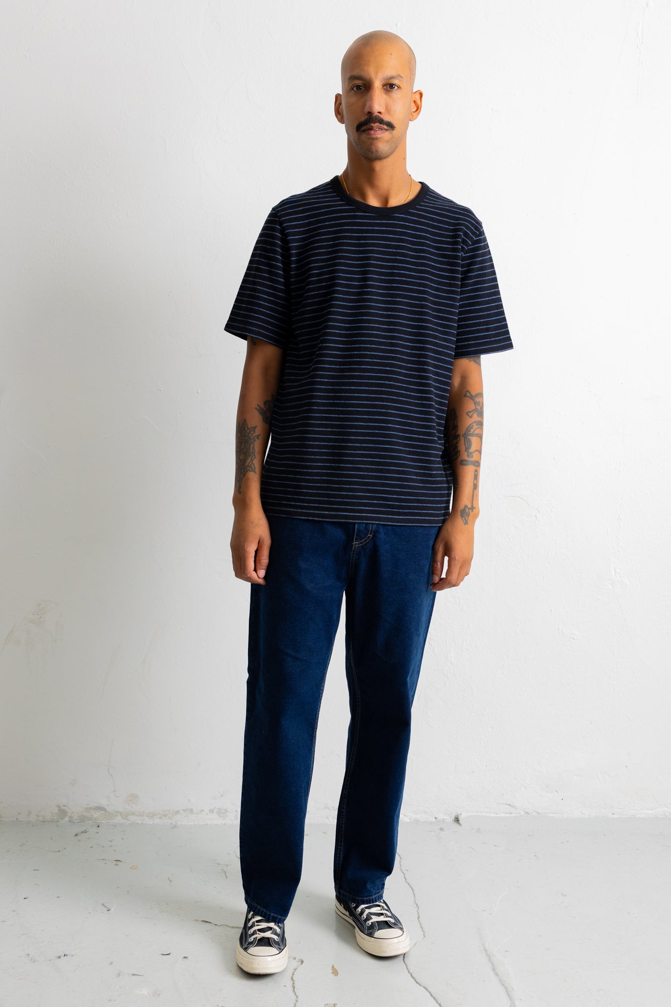 A man wearing a black short-sleeve t-shirt with thin horizontal blue stripes, dark blue jeans, and black-and-white sneakers, standing against a white studio wall.
