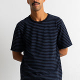 A man wearing a black short-sleeve t-shirt with thin horizontal blue stripes, paired with blue jeans, standing against a white wall.