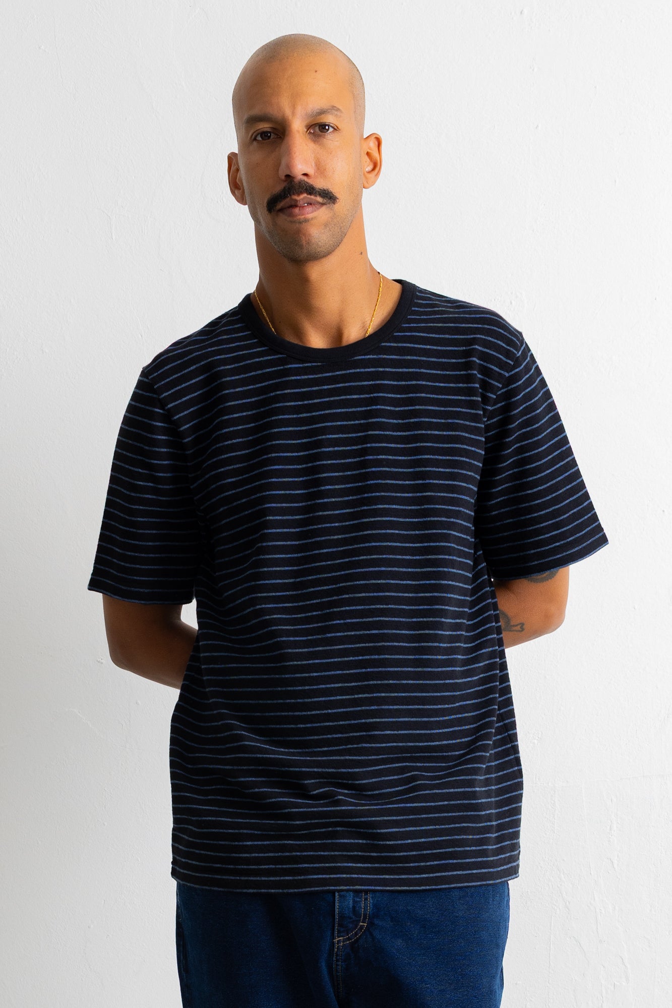 A man wearing a black short-sleeve t-shirt with thin horizontal blue stripes, paired with blue jeans, standing against a white wall.