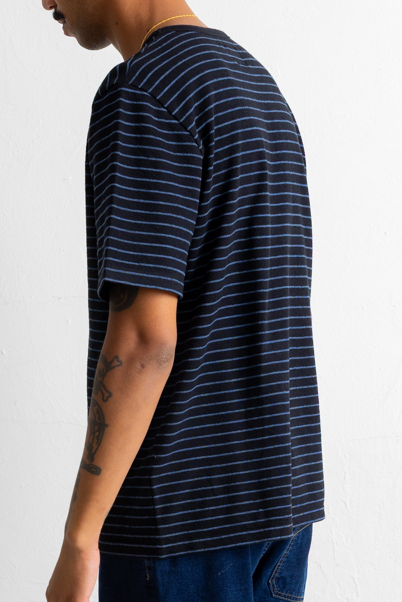 A man wearing a black short-sleeve t-shirt with thin blue horizontal stripes, shown from a side-back angle against a plain white wall.