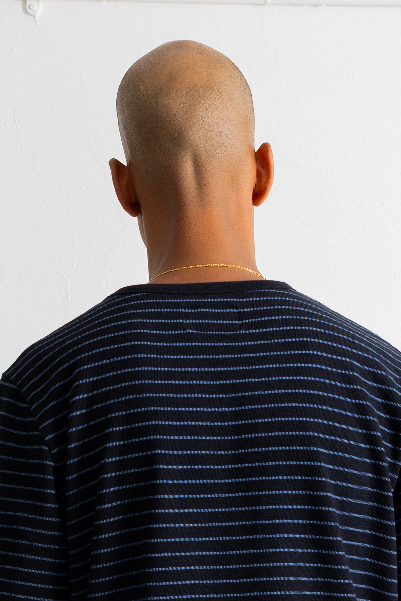 Back view of a bald man wearing a black and blue striped t-shirt, standing against a plain white wall.