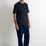 A man wearing a black and blue striped t-shirt, dark blue jeans, and black canvas sneakers, standing against a white wall.
