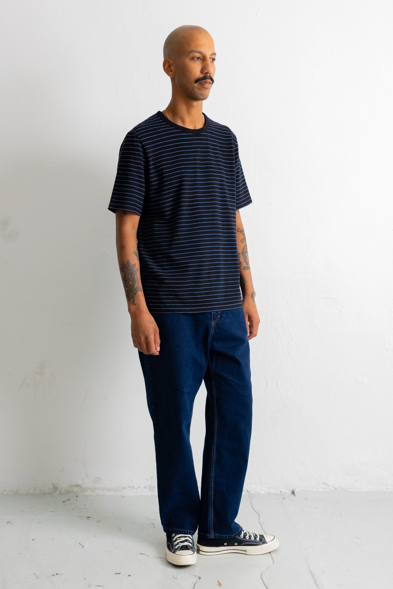 A man wearing a black and blue striped t-shirt, dark blue jeans, and black canvas sneakers, standing against a white wall.