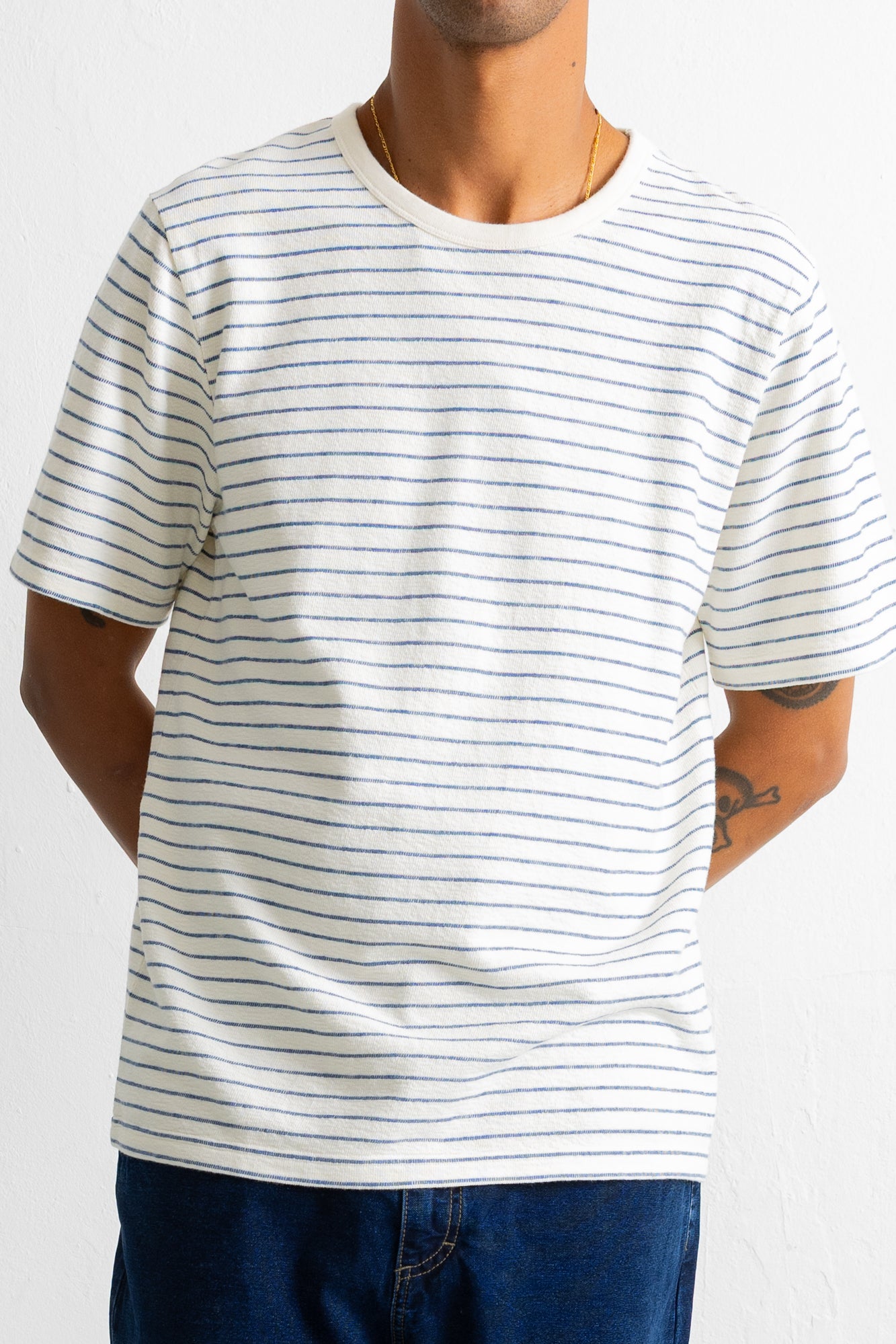 A man wearing a white short-sleeve crewneck t-shirt with thin blue horizontal stripes, paired with blue jeans, shown from the neck down against a plain white background.