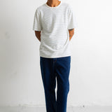 A man wearing a white short-sleeve t-shirt with thin gray horizontal stripes, dark blue jeans, and black-and-white sneakers, standing against a plain white wall.