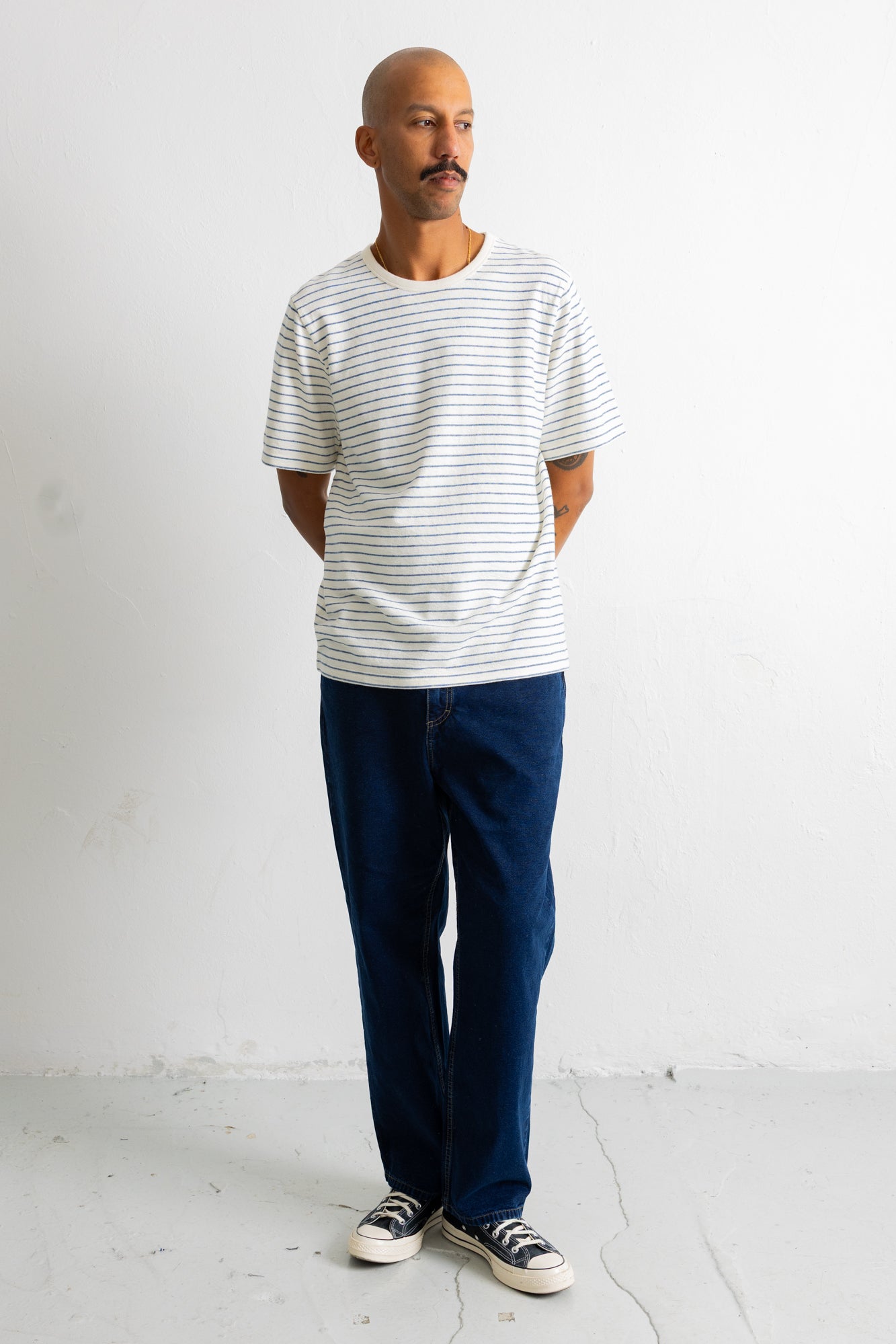 A man wearing a white short-sleeve t-shirt with thin gray horizontal stripes, dark blue jeans, and black-and-white sneakers, standing against a plain white wall.