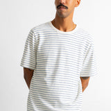 A man wearing a white short-sleeve crewneck t-shirt with thin blue horizontal stripes, paired with dark blue jeans, standing against a plain white wall.
