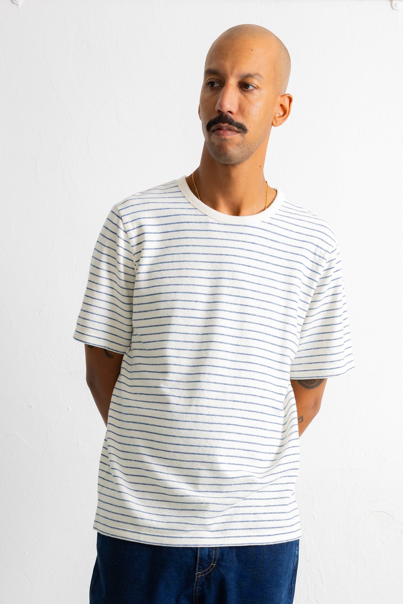 A man wearing a white short-sleeve crewneck t-shirt with thin blue horizontal stripes, paired with dark blue jeans, standing against a plain white wall.