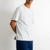 A man wearing a white short-sleeve crewneck t-shirt with thin blue horizontal stripes and dark blue jeans, standing against a plain white wall.