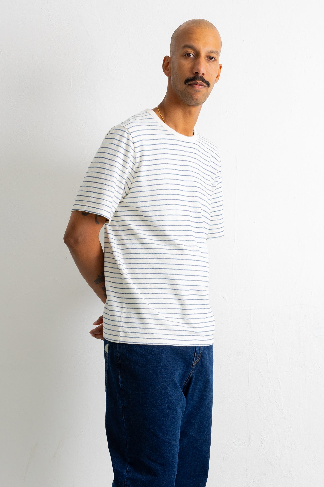 A man wearing a white short-sleeve crewneck t-shirt with thin blue horizontal stripes and dark blue jeans, standing against a plain white wall.