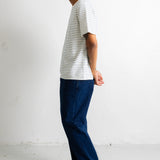 A man in profile wearing a white short-sleeve t-shirt with thin dark horizontal stripes, dark blue jeans, and black canvas sneakers, standing against a plain white wall.