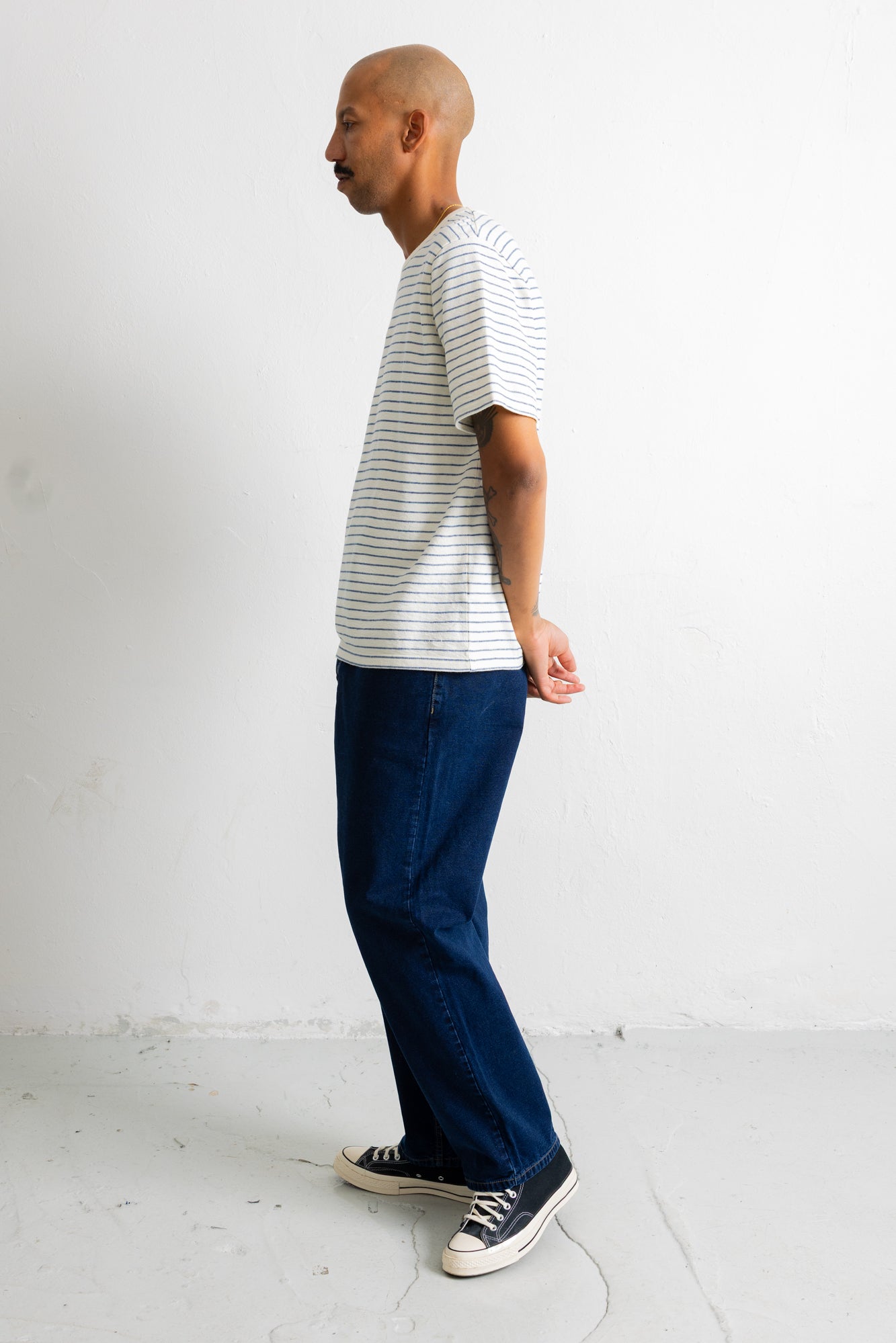A man in profile wearing a white short-sleeve t-shirt with thin dark horizontal stripes, dark blue jeans, and black canvas sneakers, standing against a plain white wall.