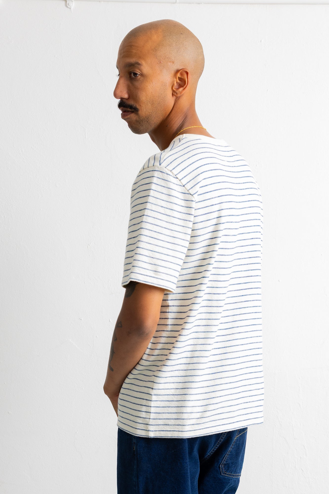 A man wearing a white short-sleeve t-shirt with thin blue horizontal stripes, shown from a side profile against a plain white wall.
