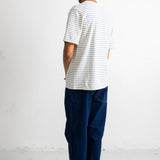 A man standing with his back to the camera wearing a white short-sleeve t-shirt with thin dark horizontal stripes, dark blue jeans, and black canvas sneakers.