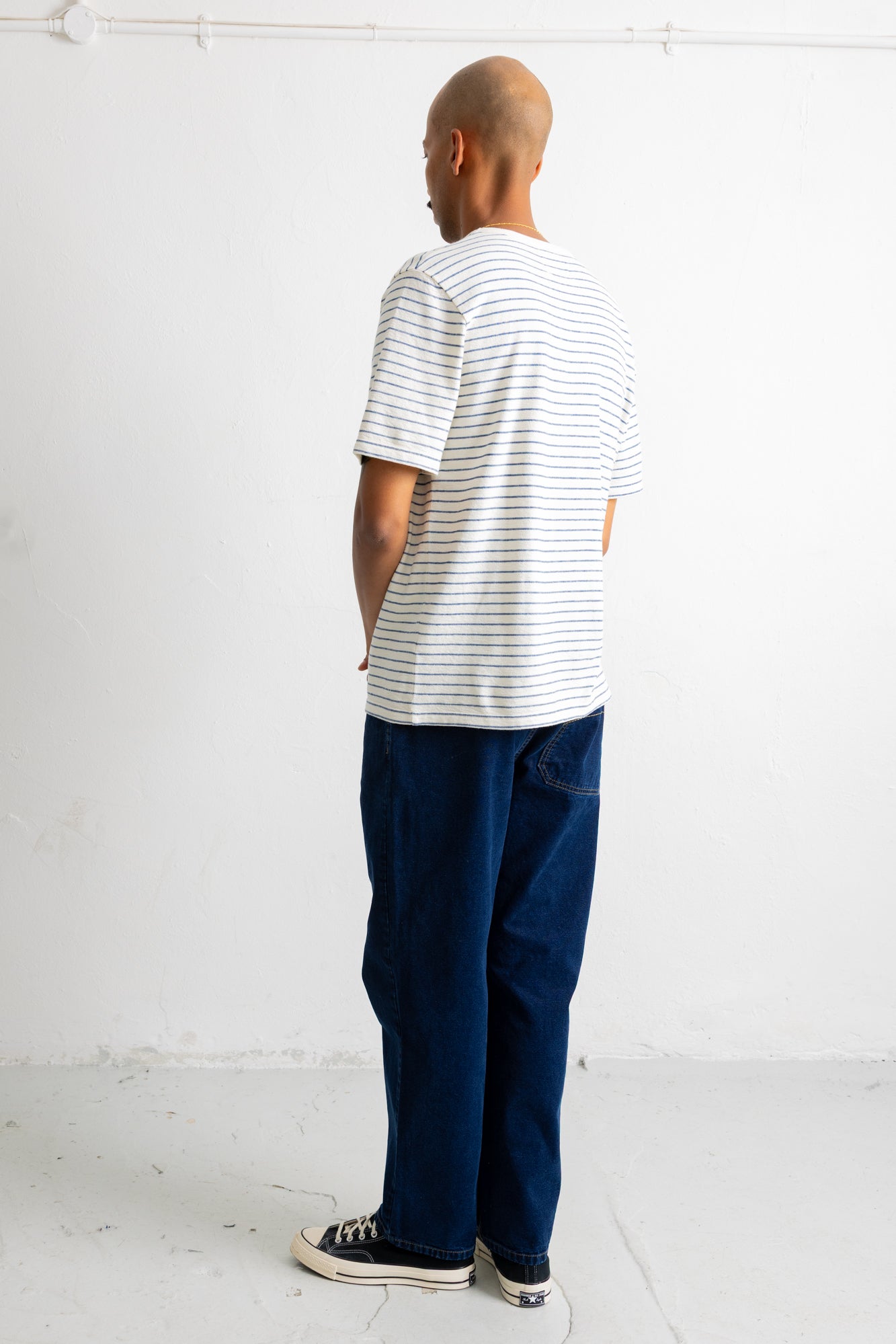 A man standing with his back to the camera wearing a white short-sleeve t-shirt with thin dark horizontal stripes, dark blue jeans, and black canvas sneakers.