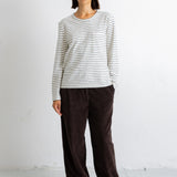 A woman stands against a white wall wearing a cream and dark gray striped long-sleeve top, dark brown wide-leg pants, and black shoes.