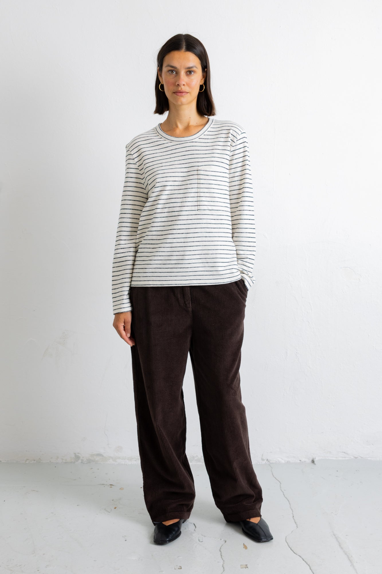 A woman stands against a white wall wearing a cream and dark gray striped long-sleeve top, dark brown wide-leg pants, and black shoes.