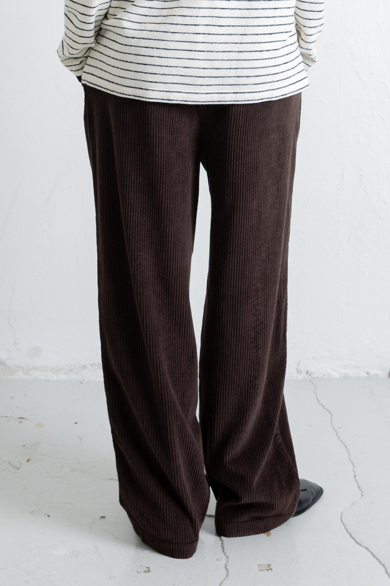 A person shown from the back wearing dark brown wide-leg corduroy pants and a cream striped long-sleeve top, standing against a white wall.